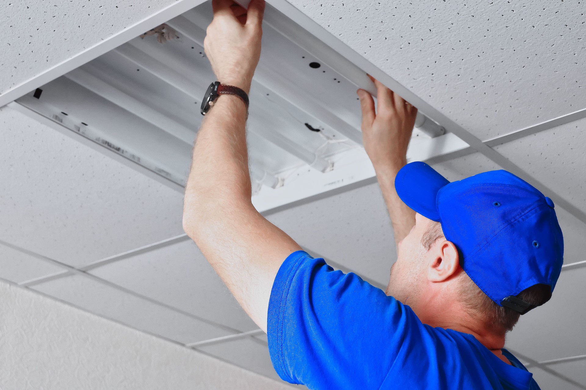 Worker repairing ceiling-mounted fluorescent light in commercial space ceiling.